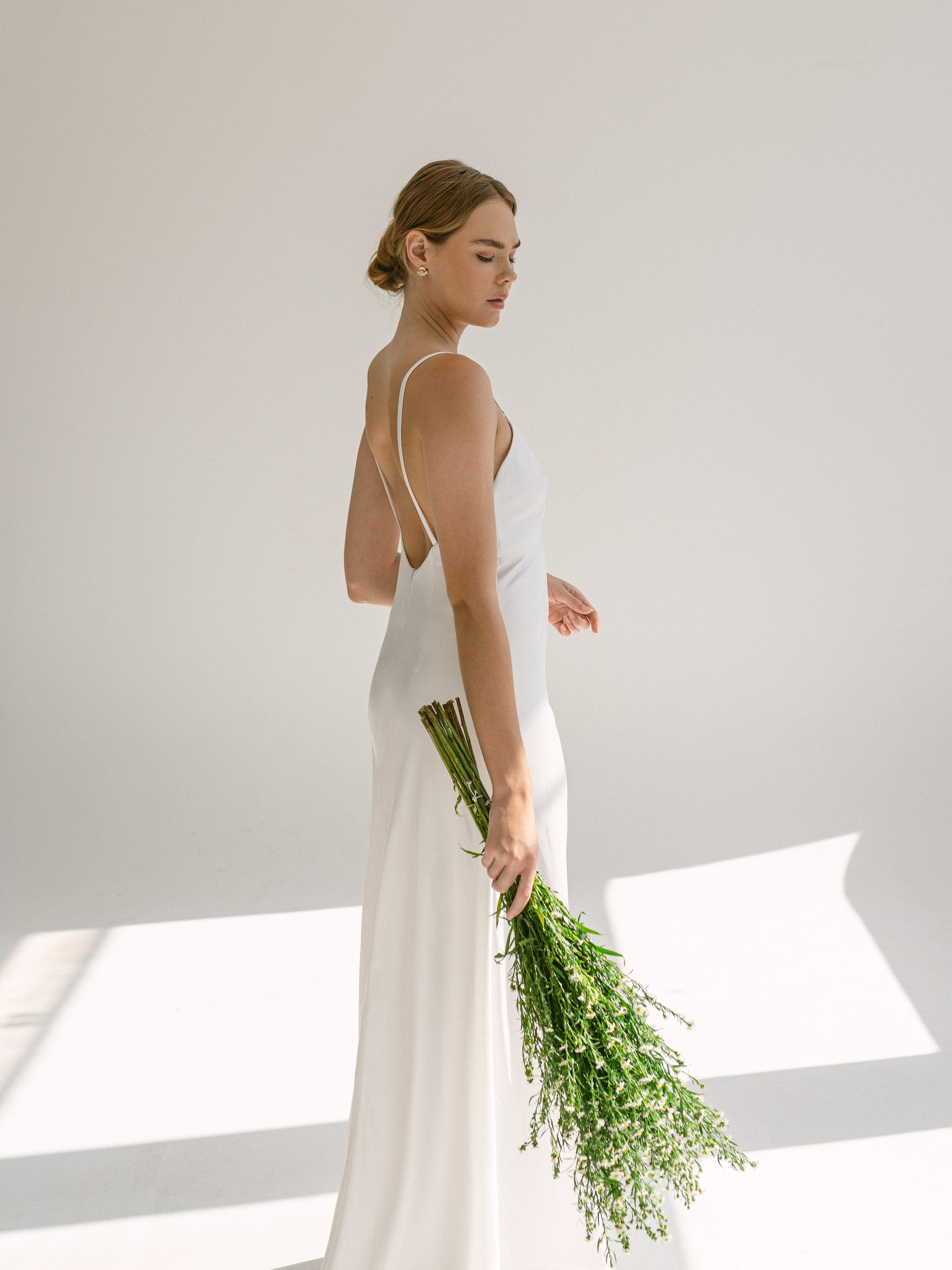 A bride standing in a white studio with a bunch of wildflower hanging by her side. 