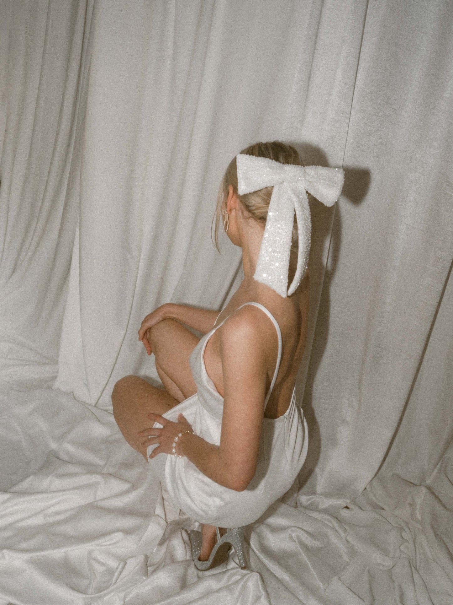 Girl in white slip dress and sparkly hair bow sitting backwards against a white satin backdrop