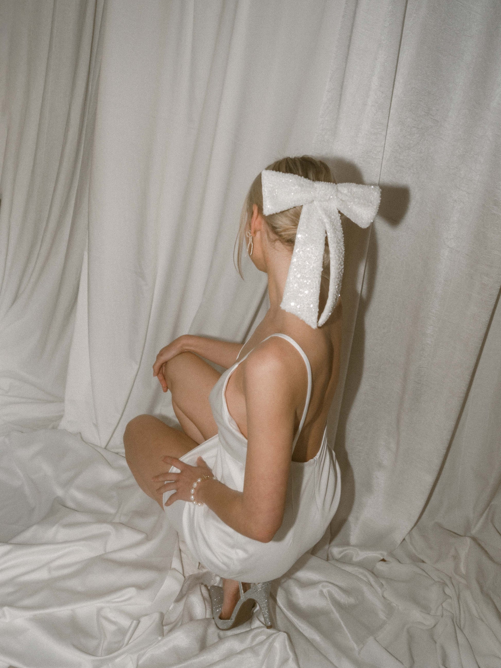 Girl in white slip dress and sparkly hair bow sitting backwards against a white satin backdrop